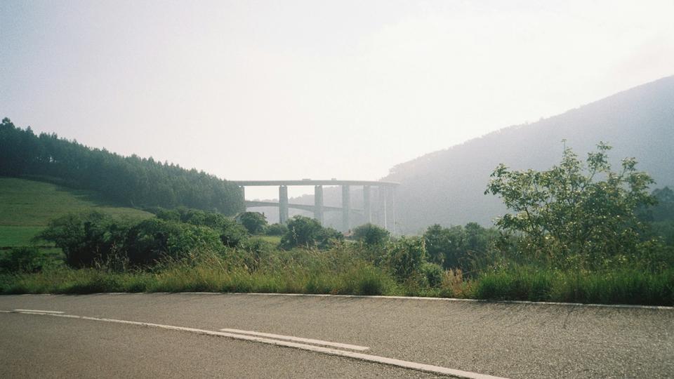 Asfaltowa droga będąca częścią Camino de Santiago w Hiszpanii, z namalowanymi liniami, dalej pas wysokiej trawy i krzewy, a w oddali wiadukt łączący zalesione wzgórza pod jasnym, lekko zamglonym niebem. Asfaltowa droga będąca częścią Camino de Santiago w Hiszpanii, z namalowanymi liniami, dalej pas wysokiej trawy i krzewy, a w oddali wiadukt łączący zalesione wzgórza pod jasnym, lekko zamglonym niebem.