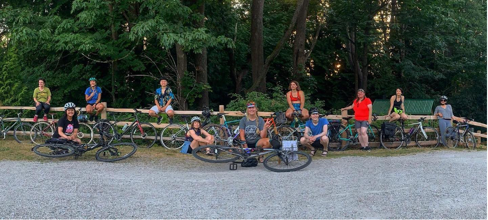 Image of a group of 11 Chill Rides members with bicycles lined up along a fence. Trees are in the background. 