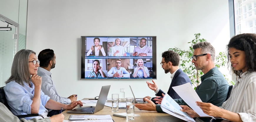 5 people in work attire sitting at a conference table with laptops and print-outs in front of them. On the wall in front of them is a screen showing 6 people on a zoom call.