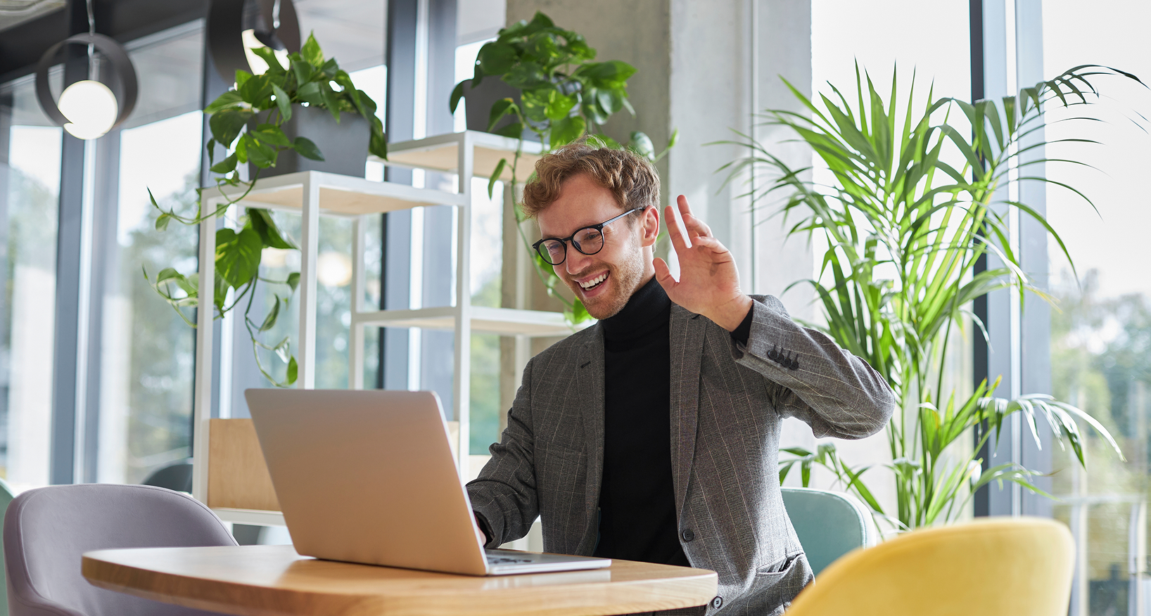 A man in business attire in a bright open office sits in front of a laptop waving to the screen.