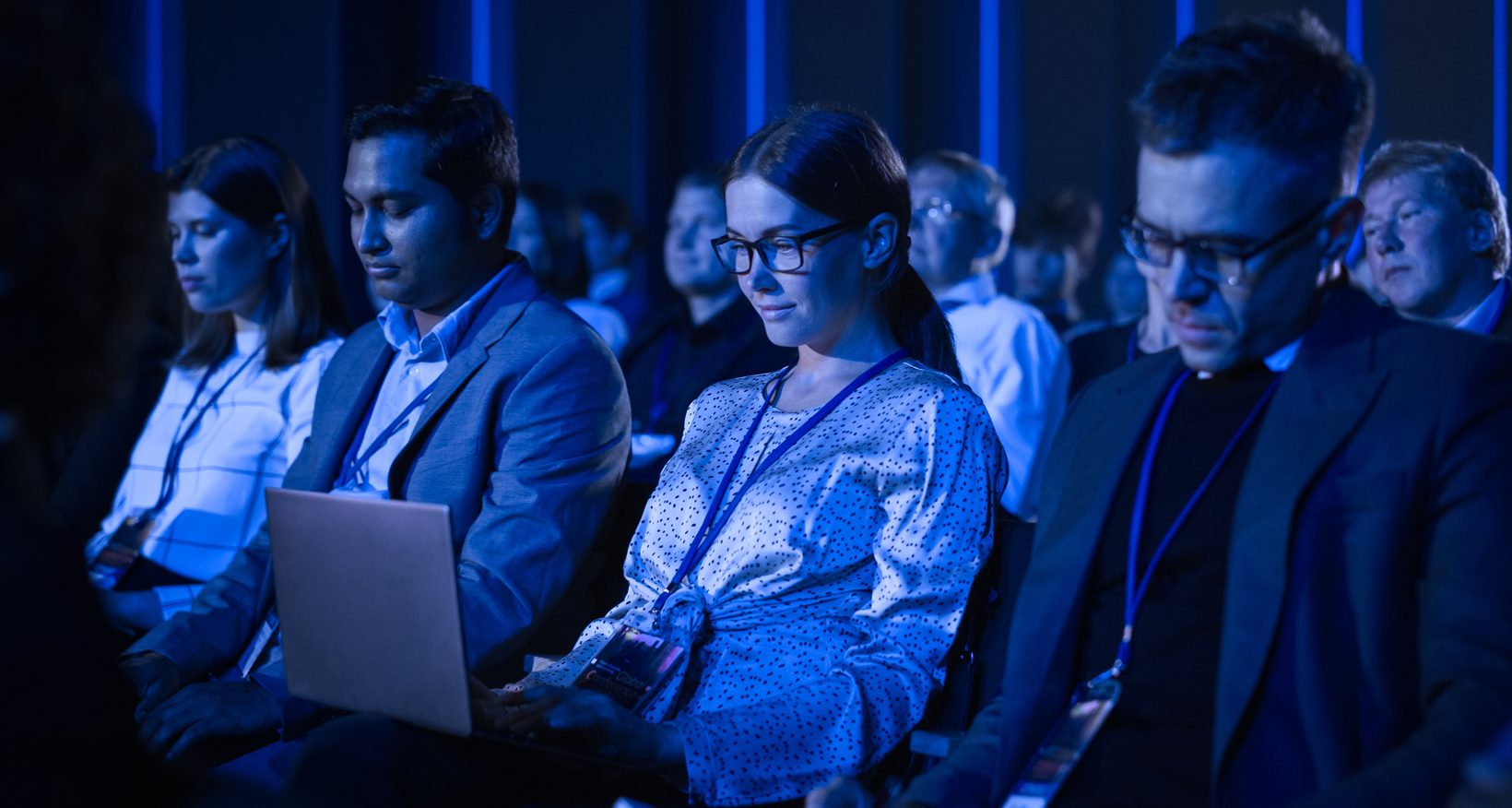 Close up shot of an audience at a conference, a audience member wearing a lanyard has her laptop on her lap. The blue light of the event filters the image.