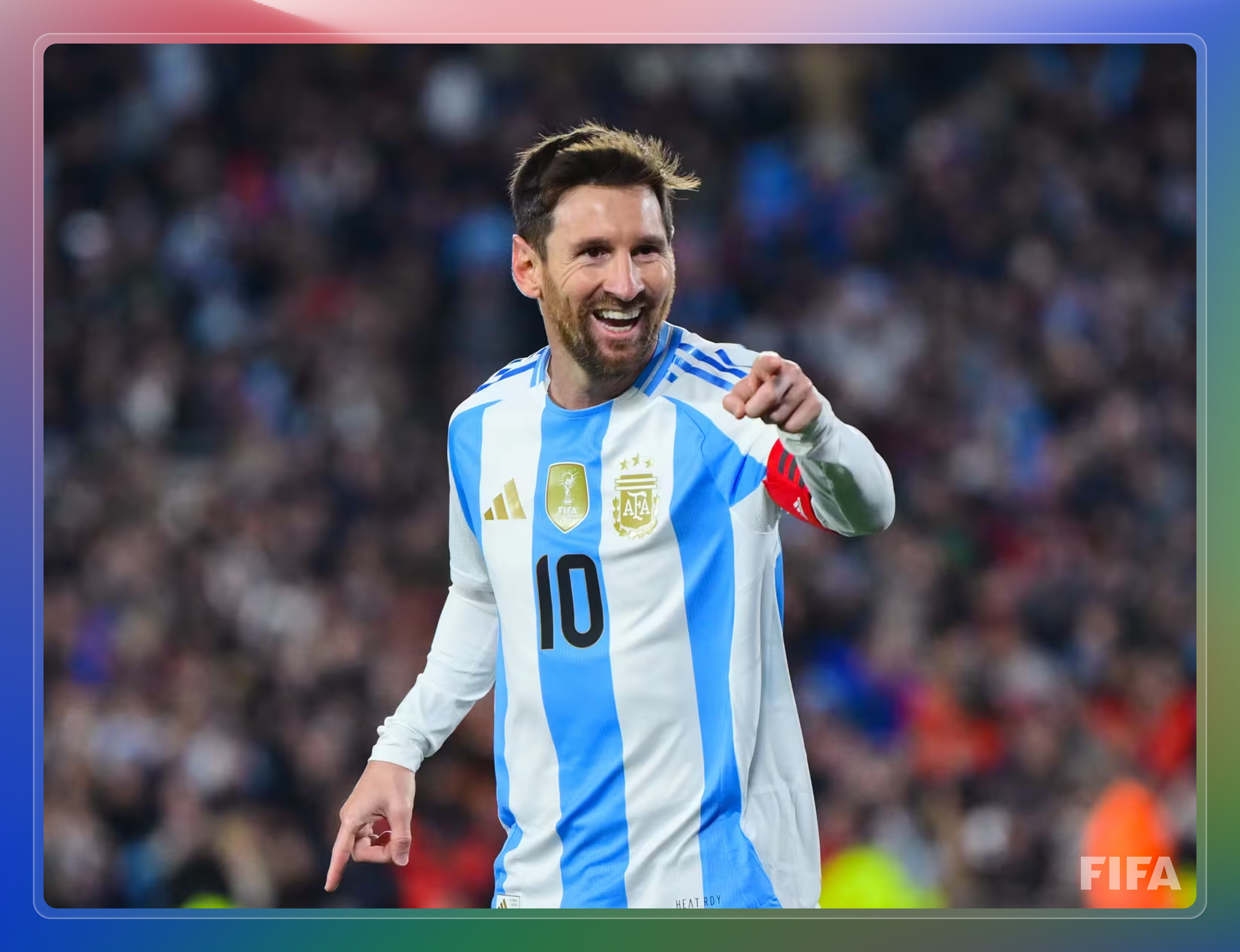A football (soccer) player in the Argentina national team kit, wearing number 10 and a captain’s armband, smiles and points toward the camera during a match. The background shows a blurred stadium crowd. The image is framed with a blue, pink, and green gradient border and features a small FIFA logo in the corner.