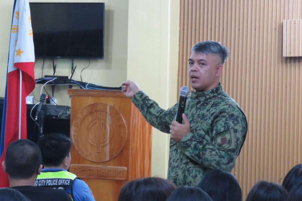 Police officer in camouflage uniform speaking at a podium with a microphone during the seminar, with a Philippine flag beside him
