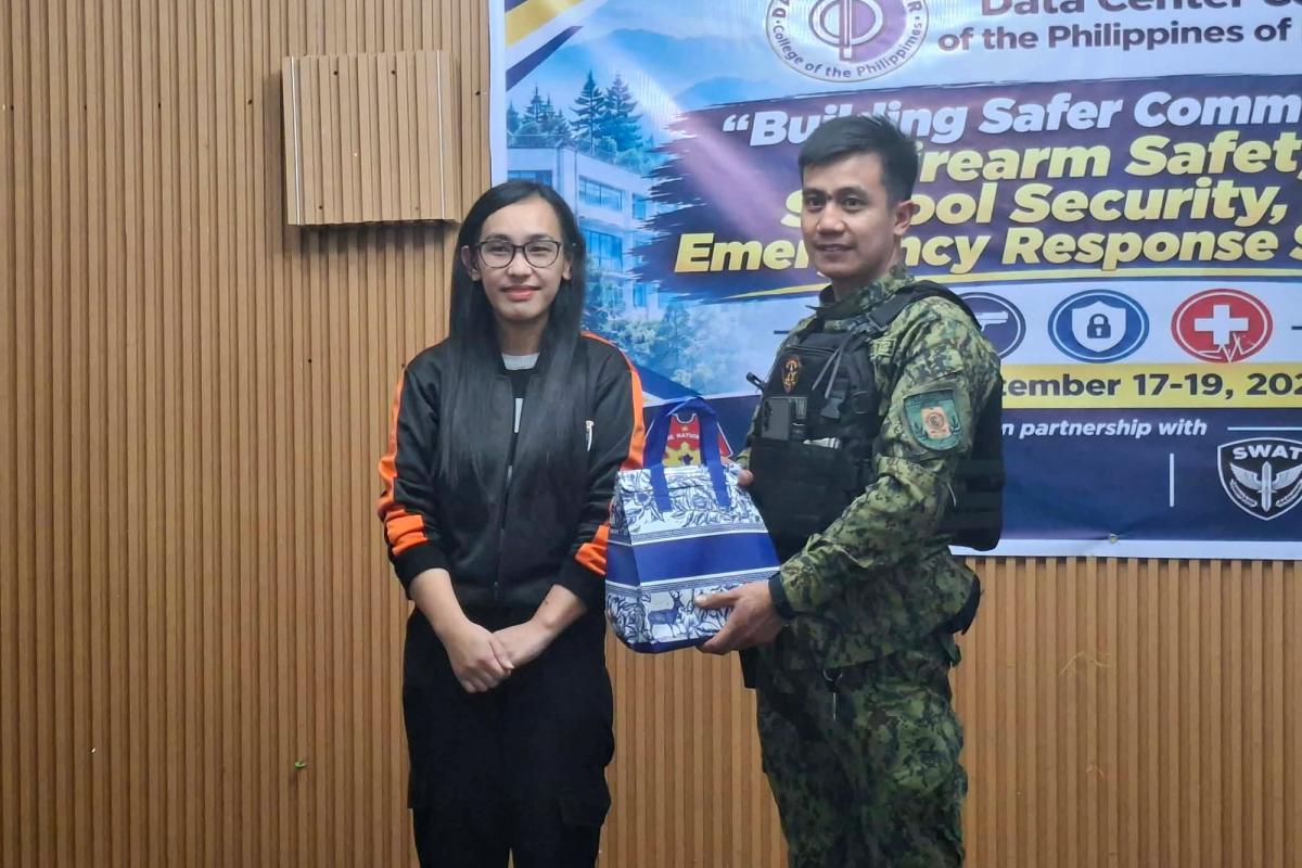 DCCP representative handing a token of appreciation to a police officer in camouflage uniform in front of the Building Safer Communities seminar banner