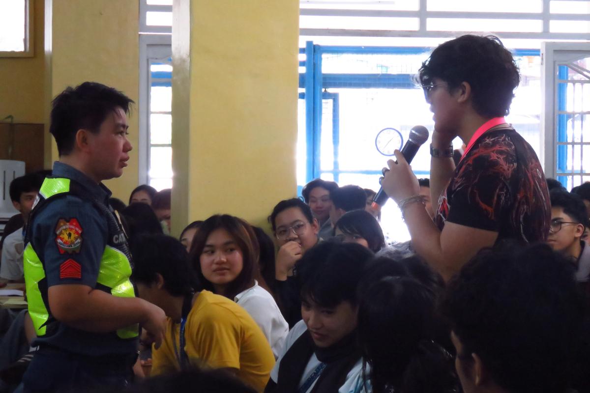 Speaker holding a microphone while addressing students in the seminar hall, with a Baguio City police officer in a reflective safety vest standing beside him