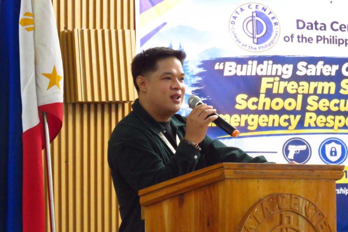 Narvey A. Sarol speaking at a wooden podium beside the Philippine flag, in front of the Building Safer Communities seminar banner