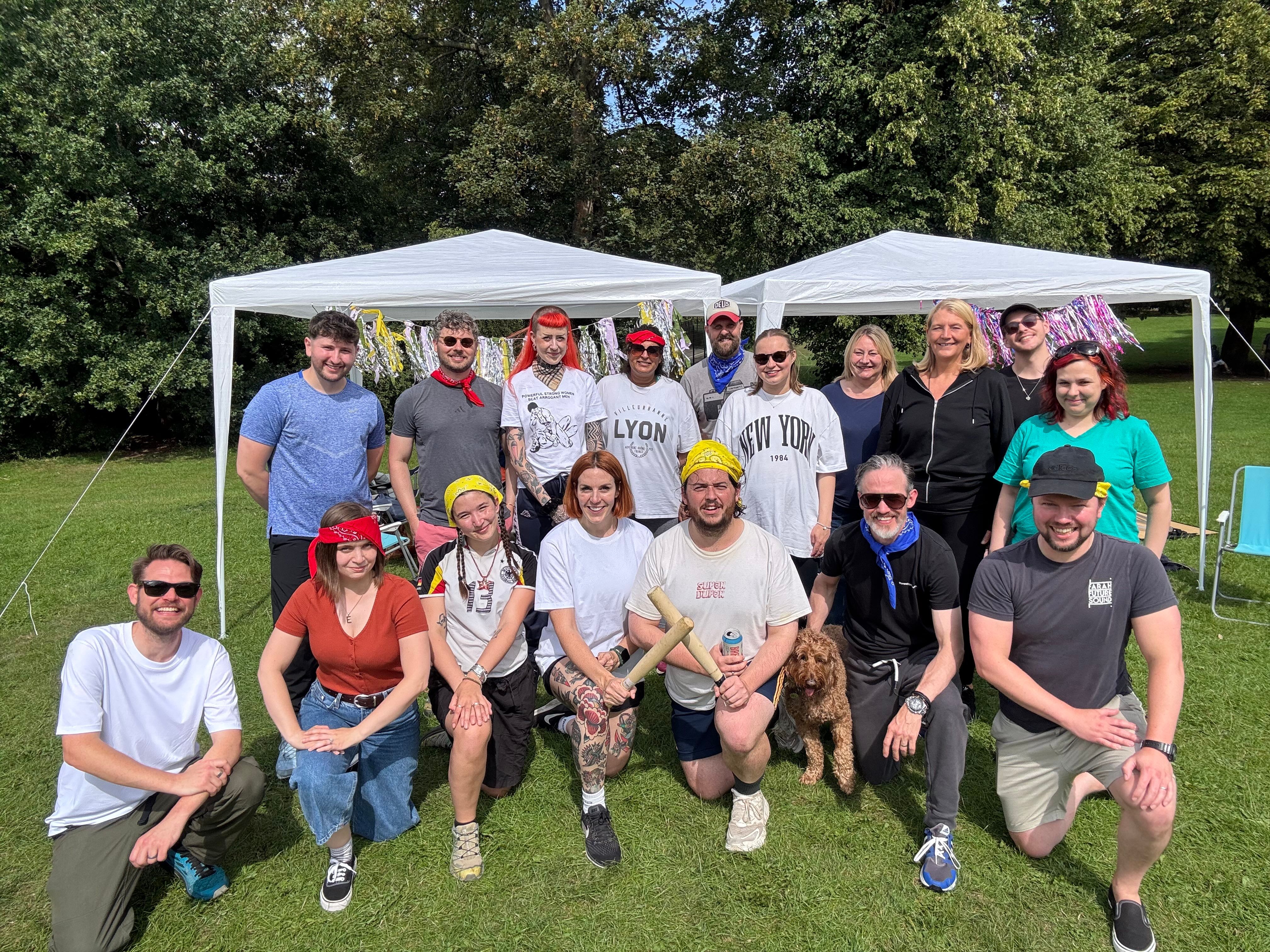 A group of around 17 people smiling and posing for a photo in a park during a team sports day. They are standing and kneeling in front of two white gazebos decorated with streamers, with trees and grass in the background. Some wear colourful bandanas, and one person holds a dog.