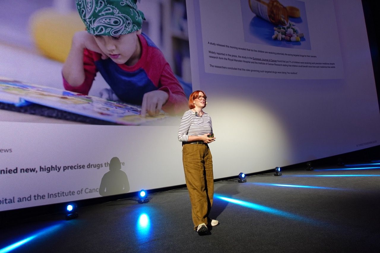 Woman presents on very large stage. She's white, 40 years old with a ginger bob, stripy top, loose trousers and trainers. She's looking relaxed, with the bottom on the screen showing behind her. It looks like a massive auditorium but we can't see.