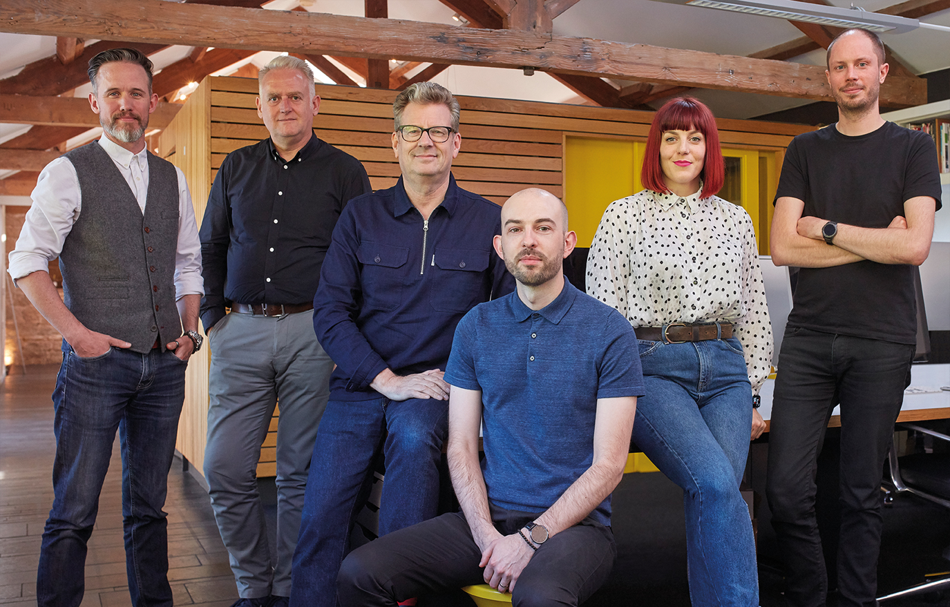 Five men and one woman sit in the middle of a creative studio, looking at the camera