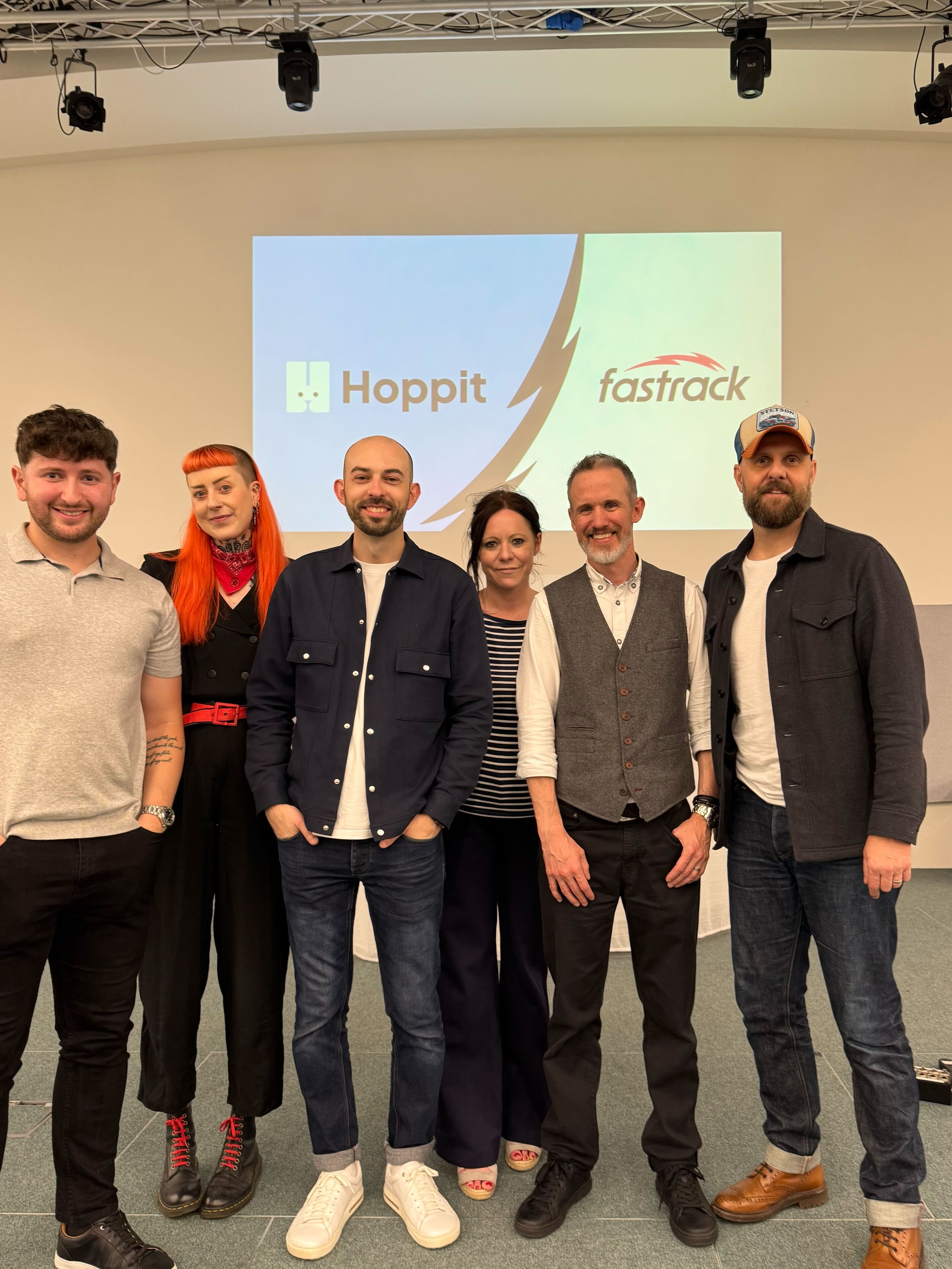 A group of 6 individuals - 4 men and 2 women, smiling for a photograph in front of a screen that says 'Hoppit' & 'Fastrack'.