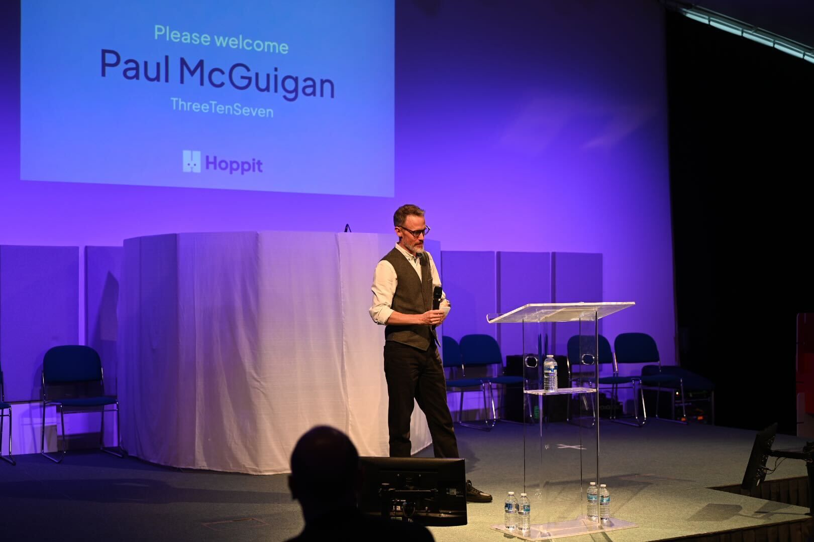 A man on stage with a purple lit background, with the text 'Please welcome Paul McGuigan' on the screen behind him.