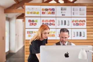 Studious looking gentleman sits at a Mac, a younger woman leaning over his shoulder. It's a creative studio in the background