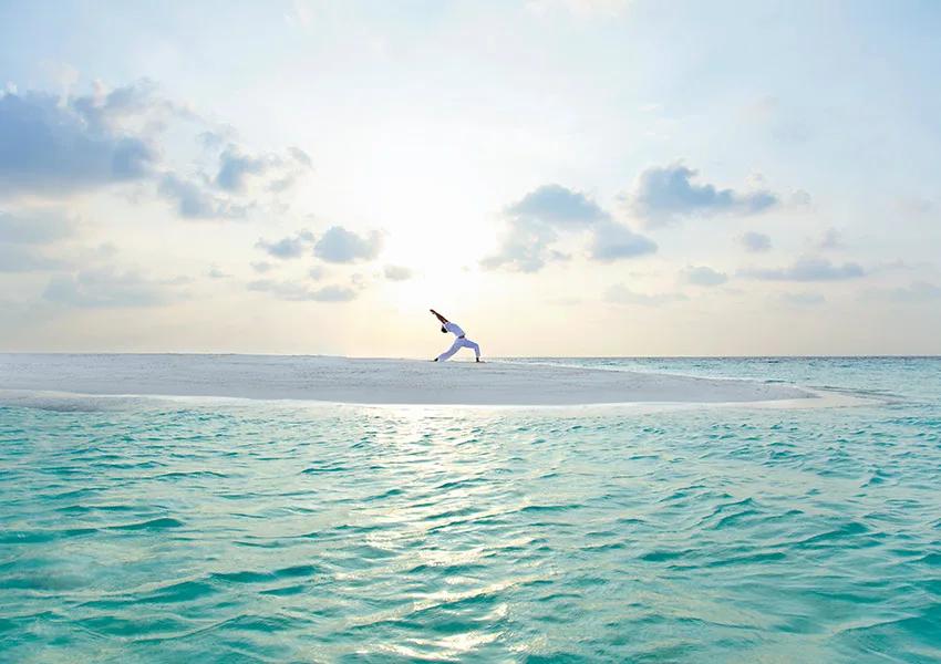 A guest in a yoga warrior pose on a narrow sandbank surrounded by shallow turquoise water at sunrise