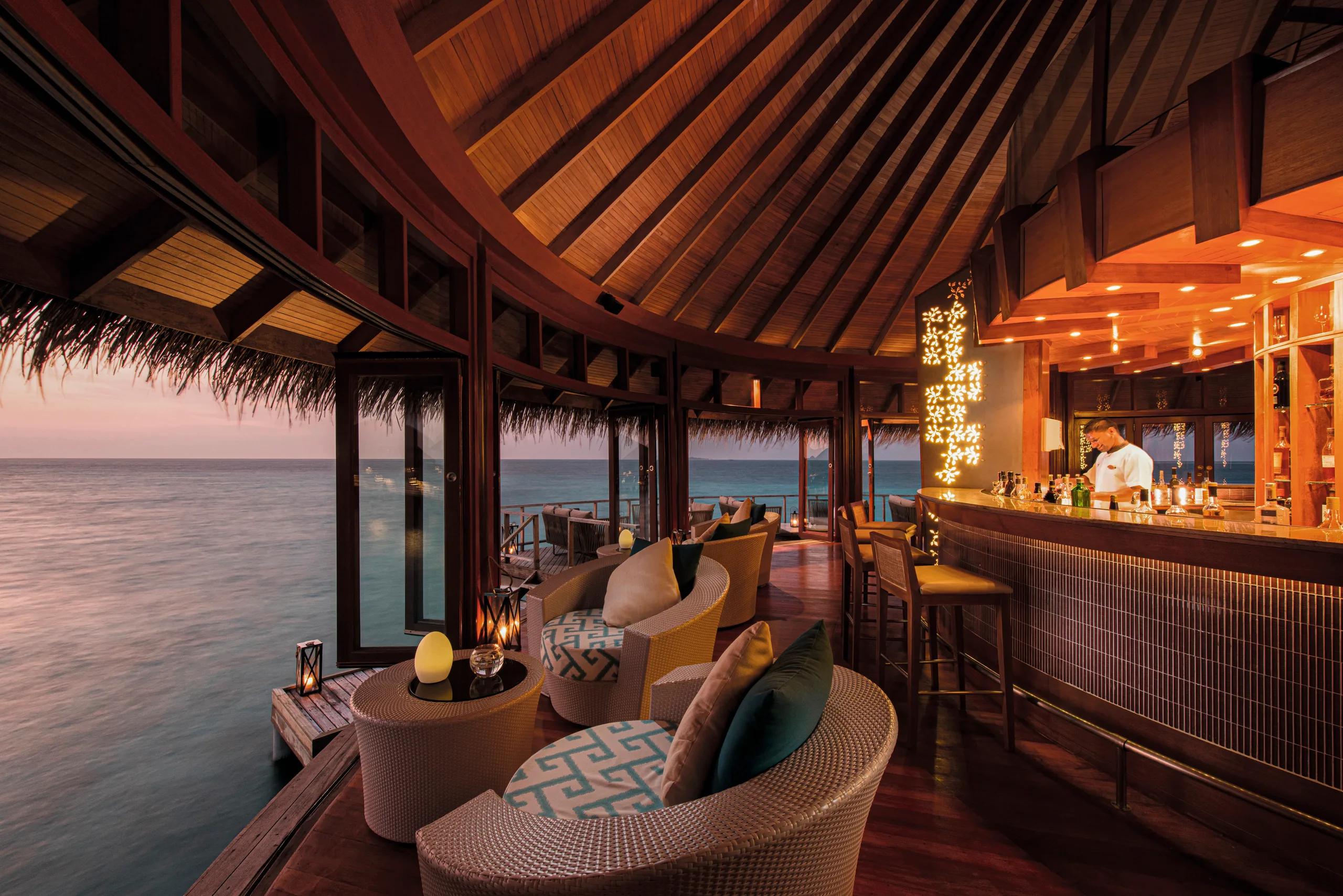 Jing Bar at dusk, rattan cocoon chairs and a lit bar counter on an overwater deck, a bartender preparing a drink, the sea visible through open doors