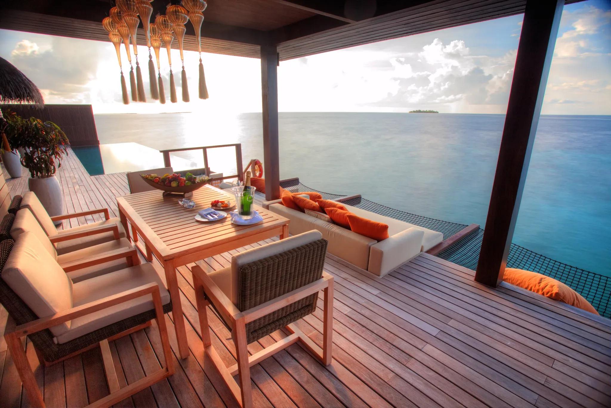 The Royal Ocean Suite's overwater dining deck at sunset, a hanging lantern chandelier above the table and open ocean beyond the netting