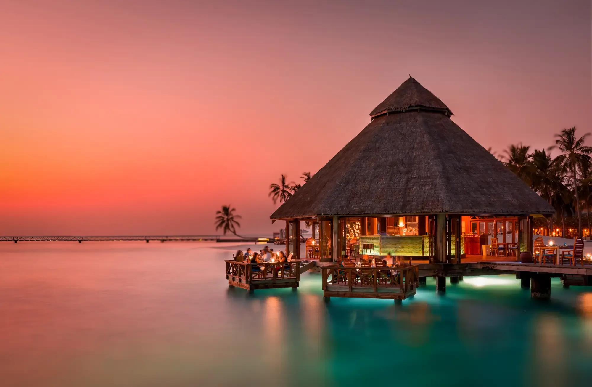 Sunset Grill's thatched-roof pavilion lit at dusk over the lagoon at Conrad Maldives Rangali