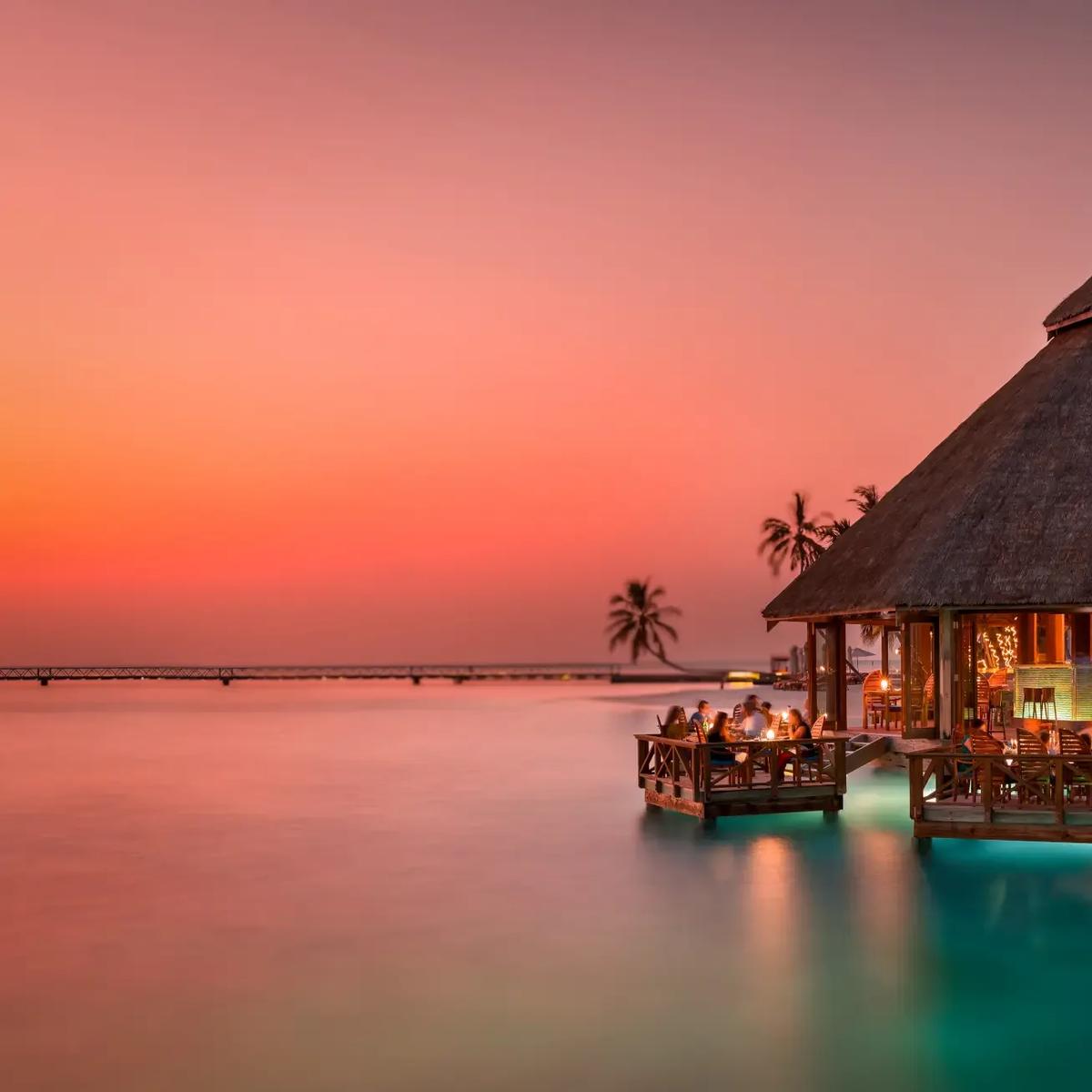 Sunset Grill's thatched-roof pavilion lit at dusk over the lagoon at Conrad Maldives Rangali