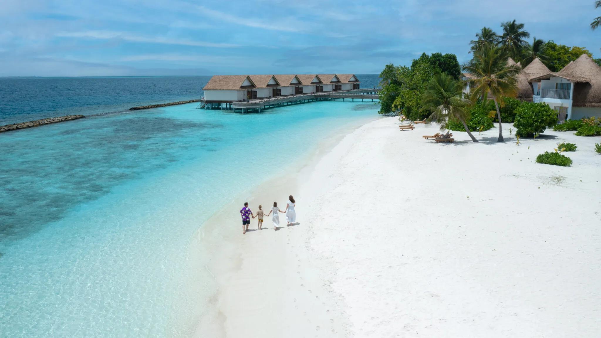 A family of four walking barefoot along Bandos's beach at low tide, the resort's overwater villas and jetty visible across the lagoon behind them