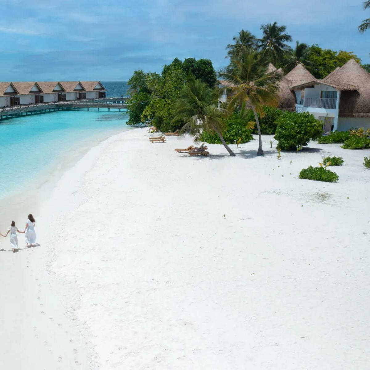 A family of four walking barefoot along Bandos's beach at low tide, the resort's overwater villas and jetty visible across the lagoon behind them