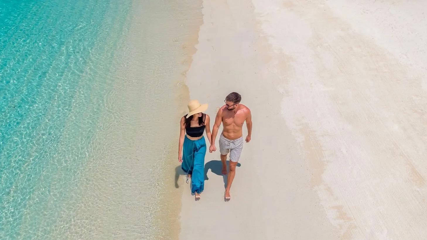 A couple walking hand in hand along the waterline between white sand and turquoise lagoon