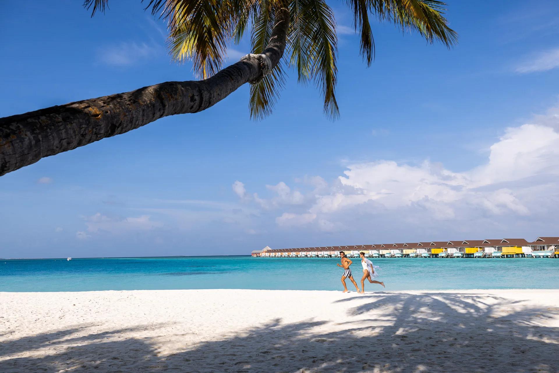 A couple jogging along the beach beneath a leaning palm tree, The Standard's overwater villa row curving into the lagoon behind them