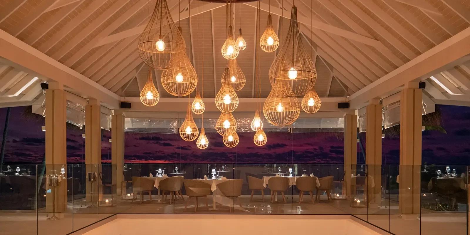 La Limonaia's open-air dining pavilion at dusk, rattan pendant lights above white-clothed tables and a darkening sky beyond