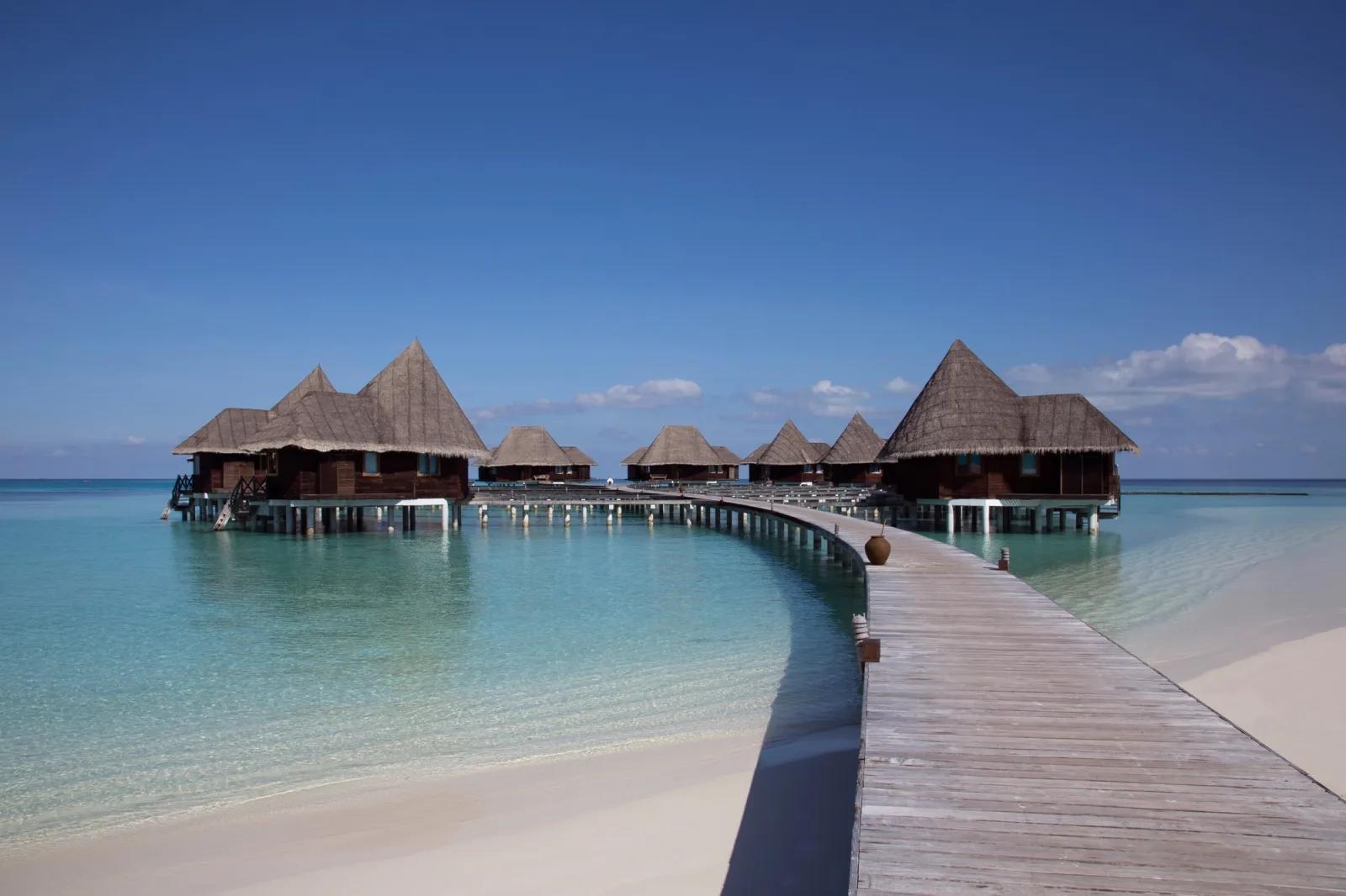 A wooden boardwalk leading out across turquoise water to Coco Palm Dhuni Kolhu's overwater Sunset Lagoon Villas under a clear blue sky