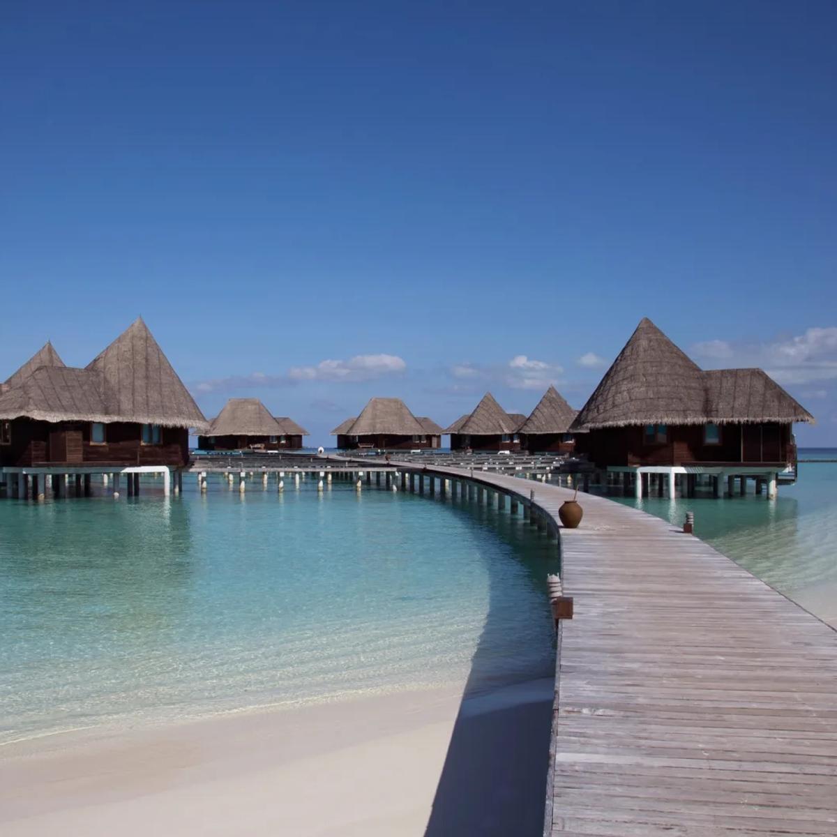 A wooden boardwalk leading out across turquoise water to Coco Palm Dhuni Kolhu's overwater Sunset Lagoon Villas under a clear blue sky