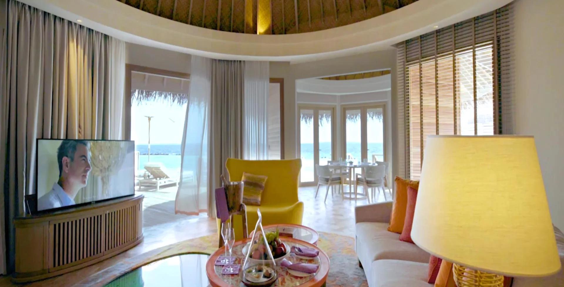 An Ocean Residence sitting room in mustard tones, with a hanging egg chair on the thatched terrace beyond and a spiral stair to one side.