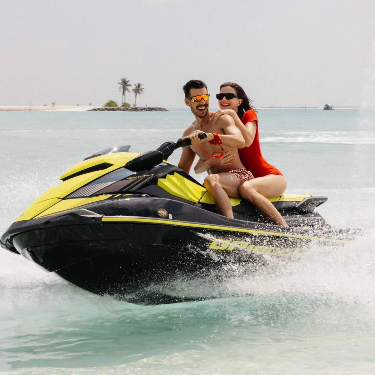 A couple riding a jet ski at speed, spray flying up behind them near a small island