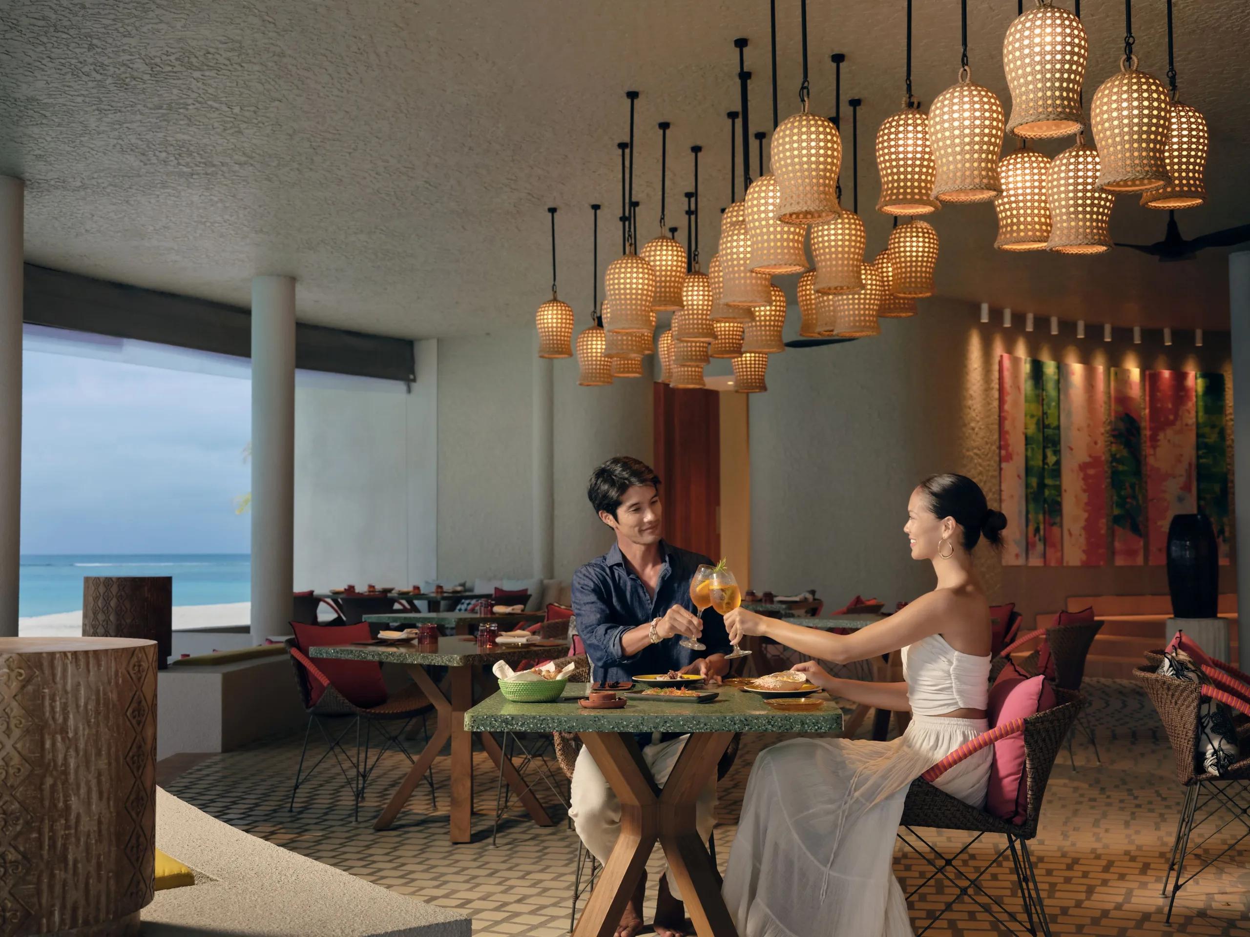 A couple toasting drinks at a table inside The Point restaurant, woven pendant lights overhead and the ocean visible beside them.