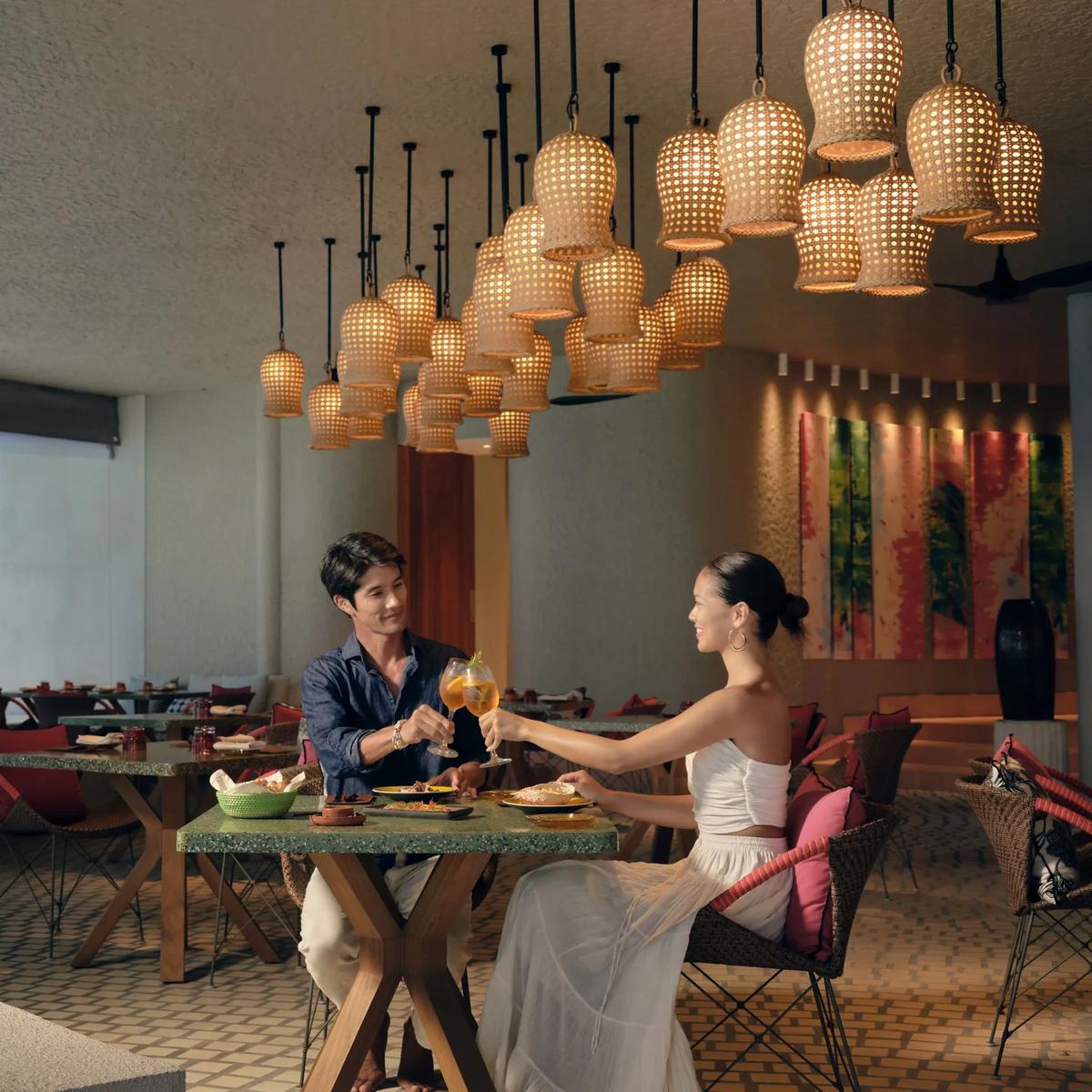 A couple toasting drinks at a table inside The Point restaurant, woven pendant lights overhead and the ocean visible beside them.