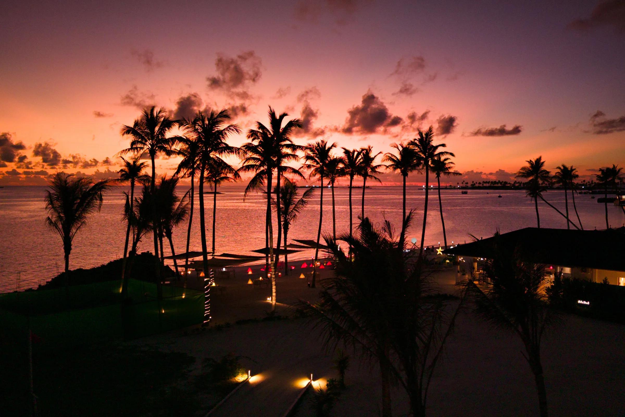 A thatched beach umbrella and loungers silhouetted against a pink sunset sky at the island's tip