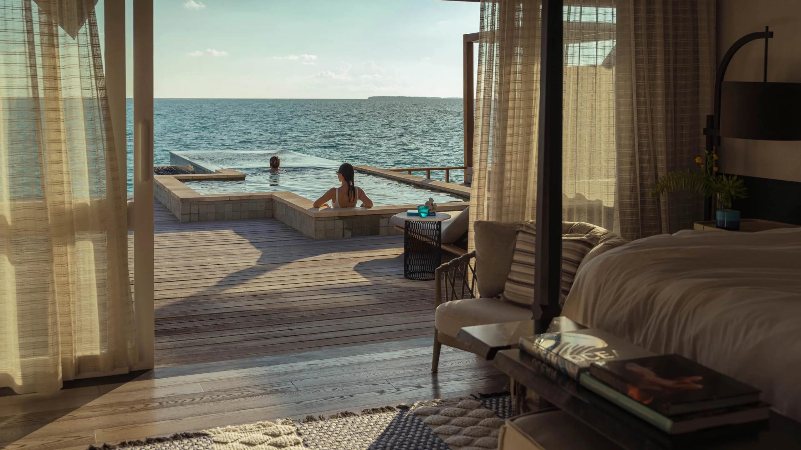 View from inside a Sunset Water Villa across its infinity-edge pool to the open ocean, two guests swimming