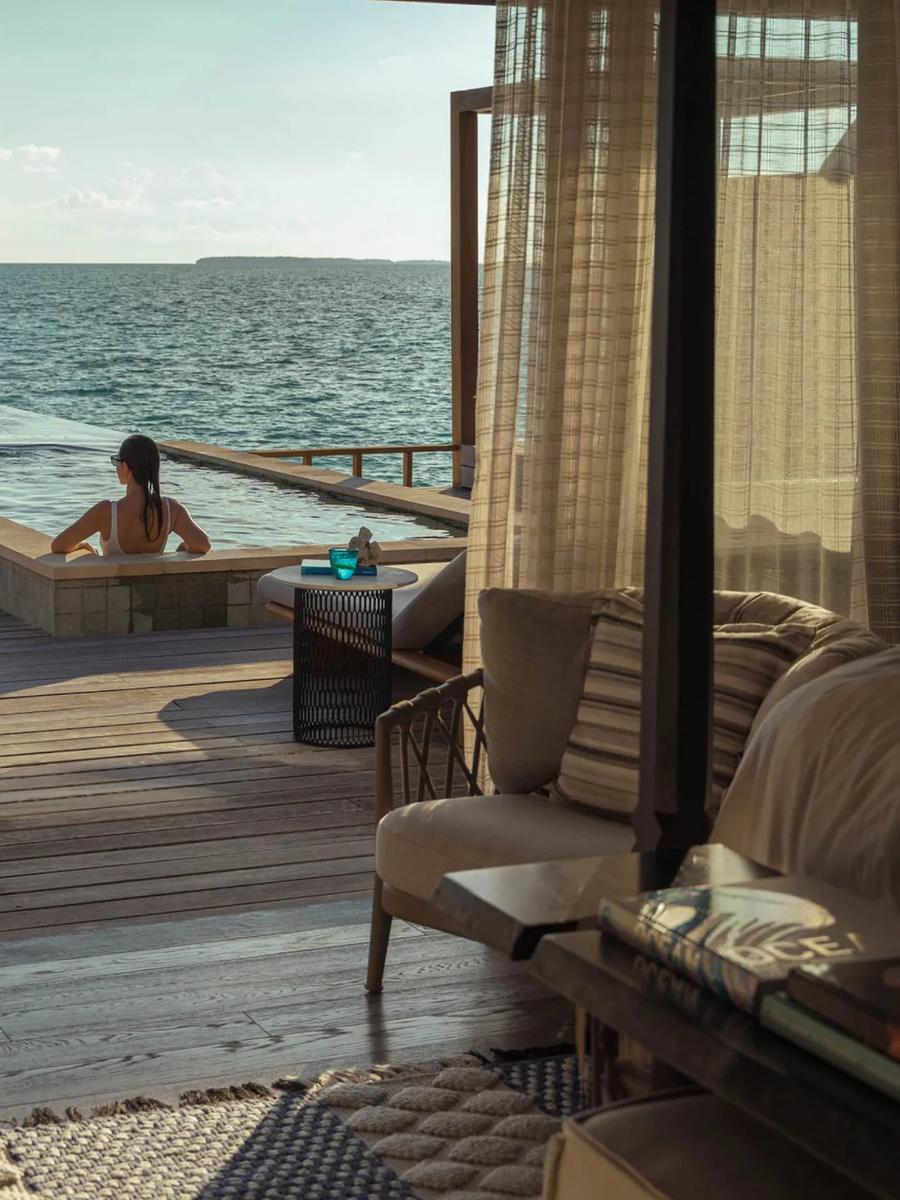 View from inside a Sunset Water Villa across its infinity-edge pool to the open ocean, two guests swimming