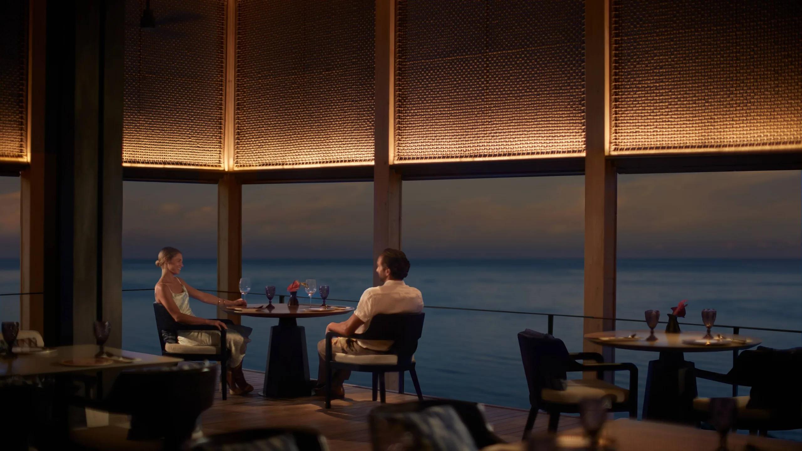 A couple dining at an overwater table as dusk settles over the ocean