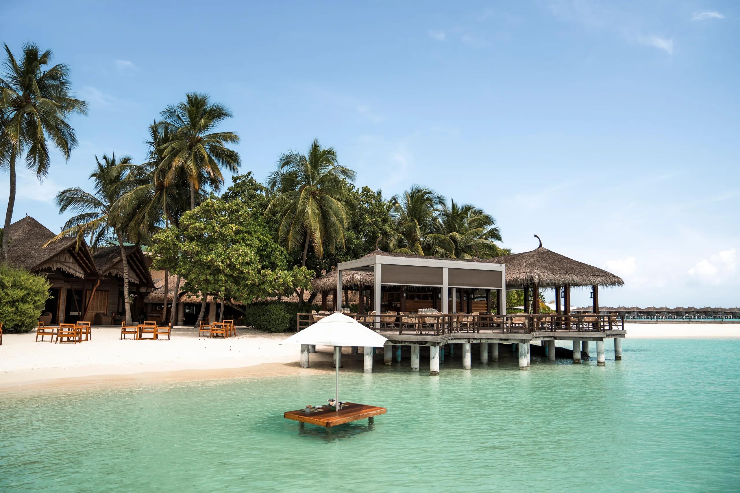 Manta Restaurant's sand-floored dining room by day, thatched roof and open sides among palm trees, beach umbrella in the lagoon