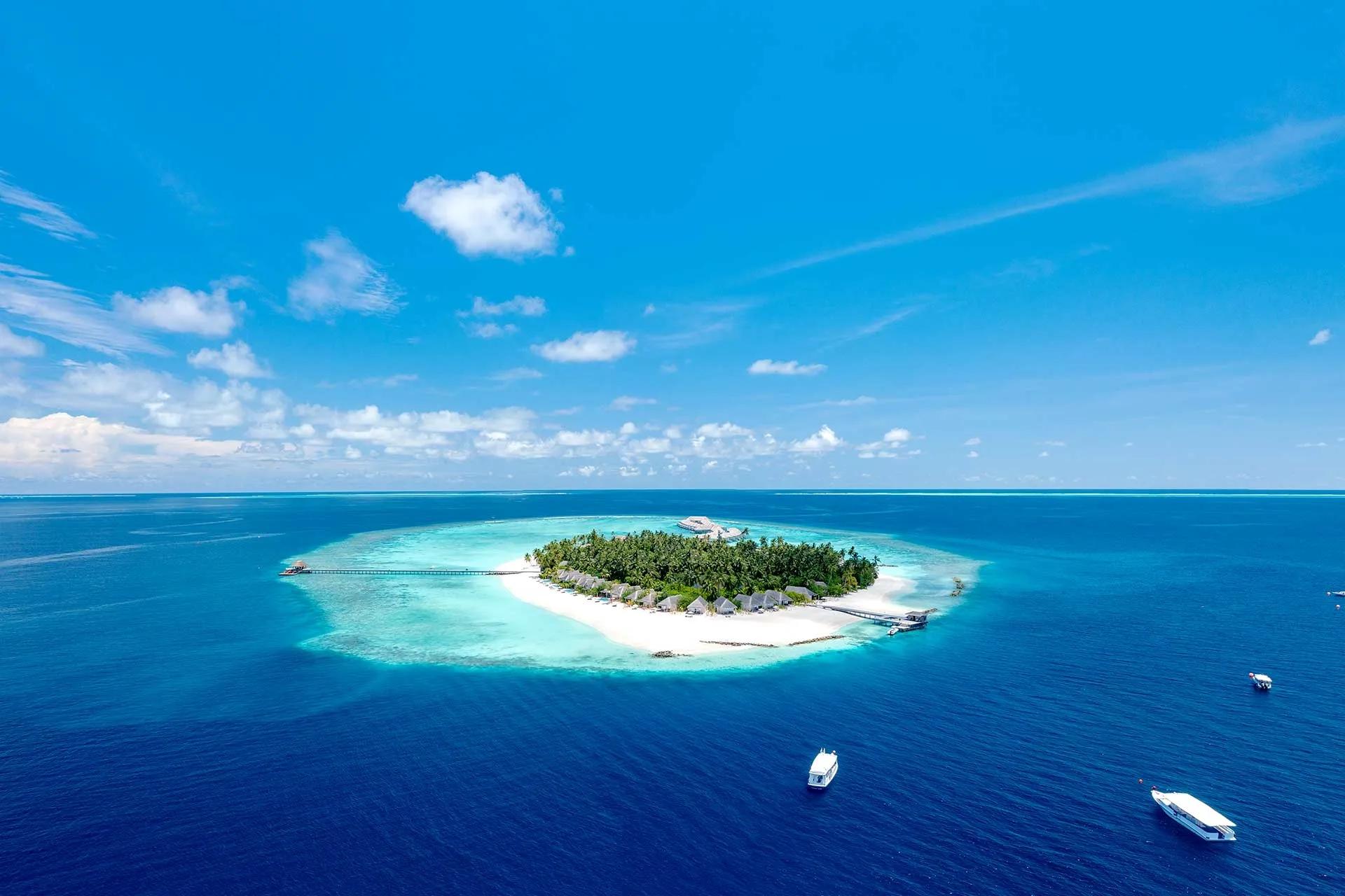 Aerial view of Maagau Island's full reef-ringed outline in Dhaalu Atoll, with two boats approaching across the outer channel