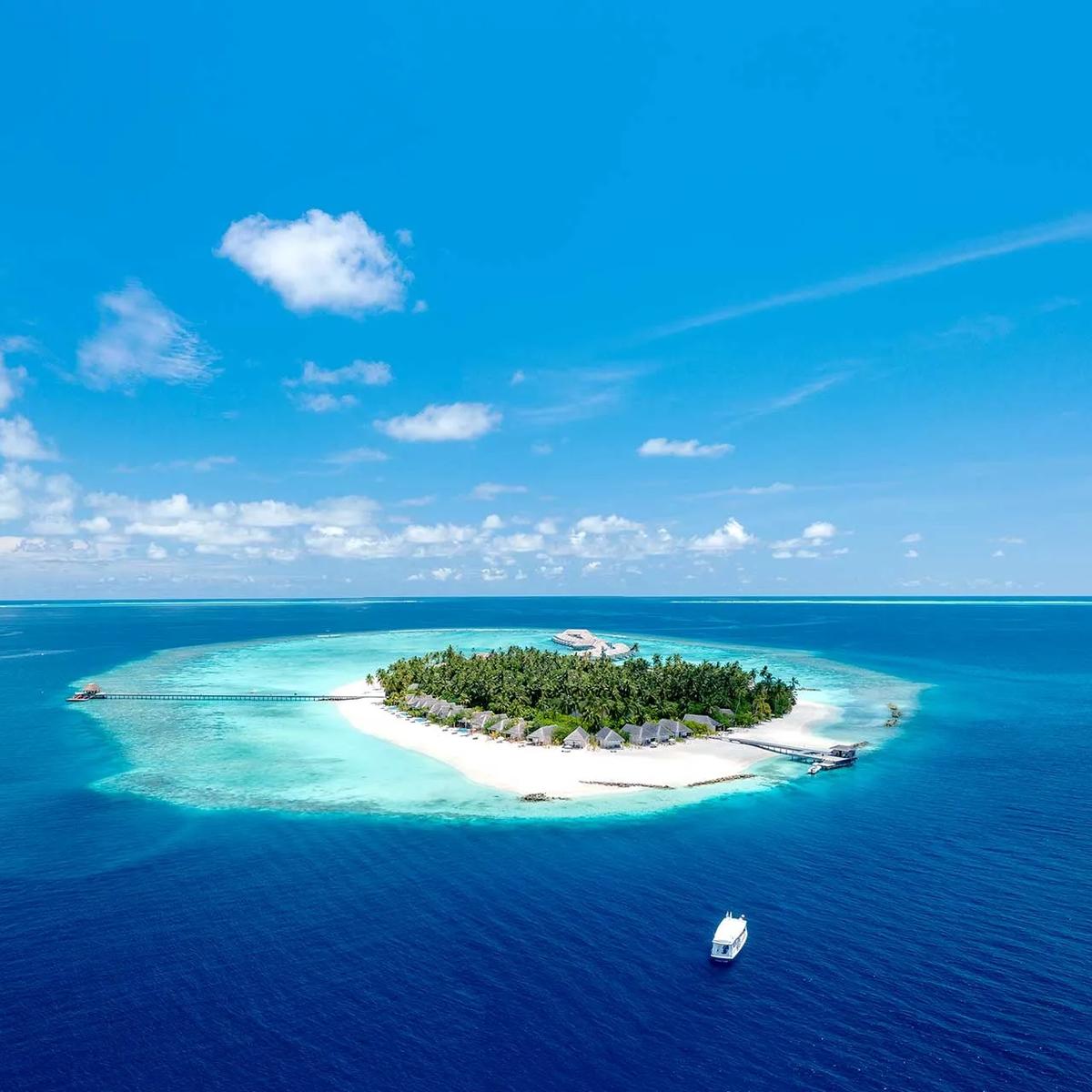 Aerial view of Maagau Island's full reef-ringed outline in Dhaalu Atoll, with two boats approaching across the outer channel