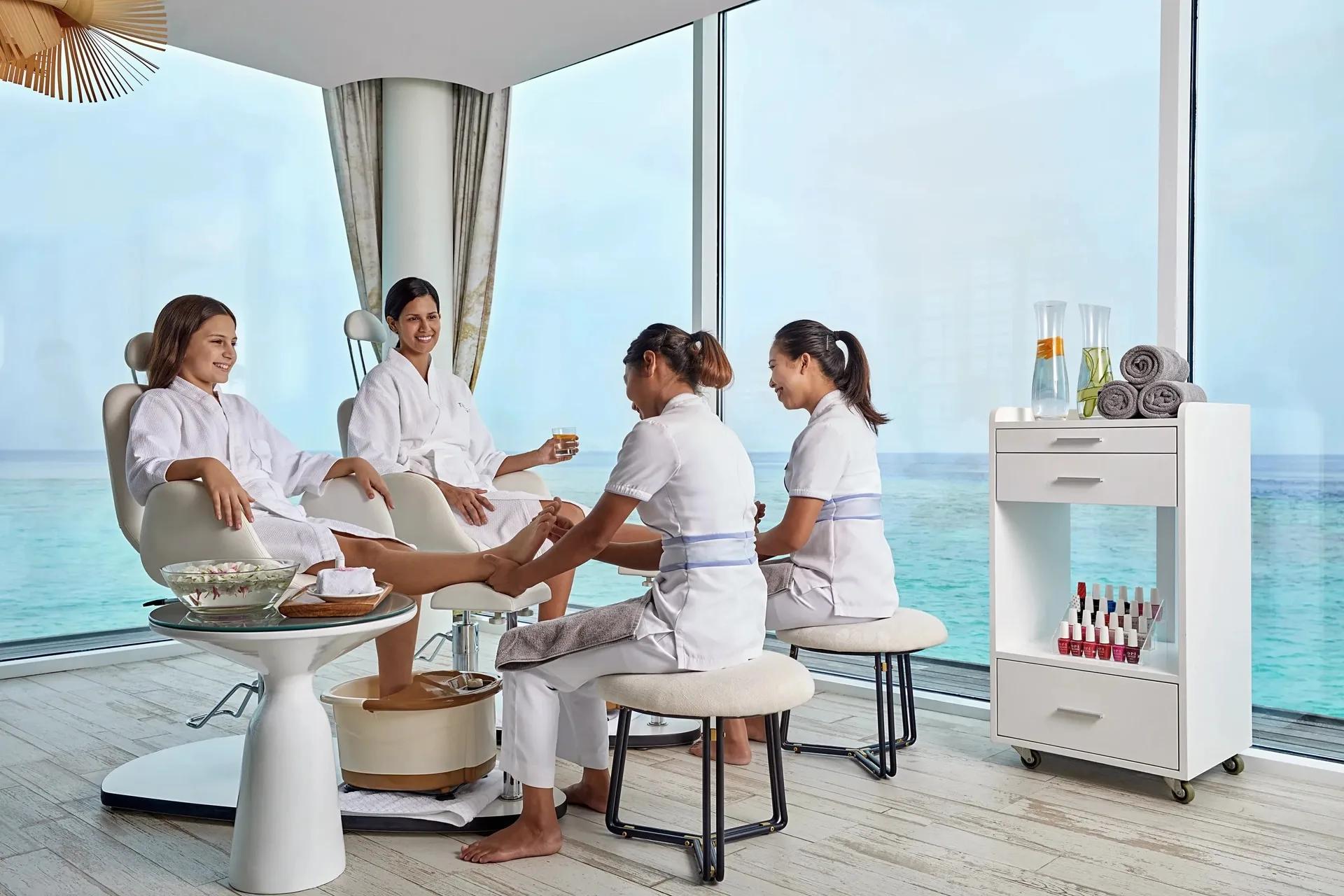 Two guests in robes receiving pedicures at an overwater beauty salon, floor-to-ceiling windows looking over the lagoon
