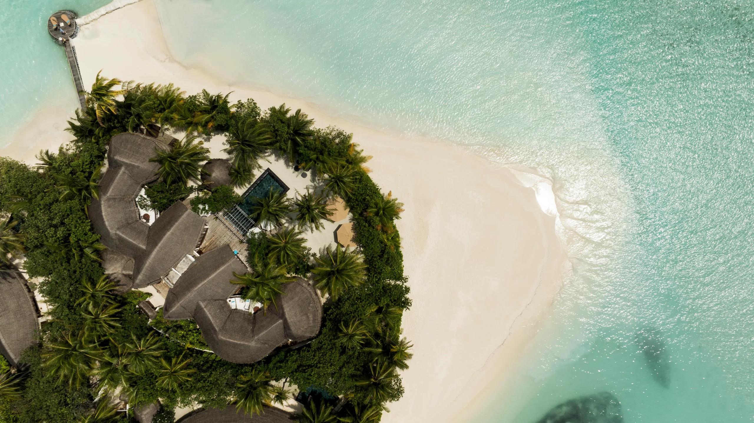 Aerial view of a cluster of thatched-roof beachfront villas with a private plunge pool between them, close to the beach and the resort's main jetty
