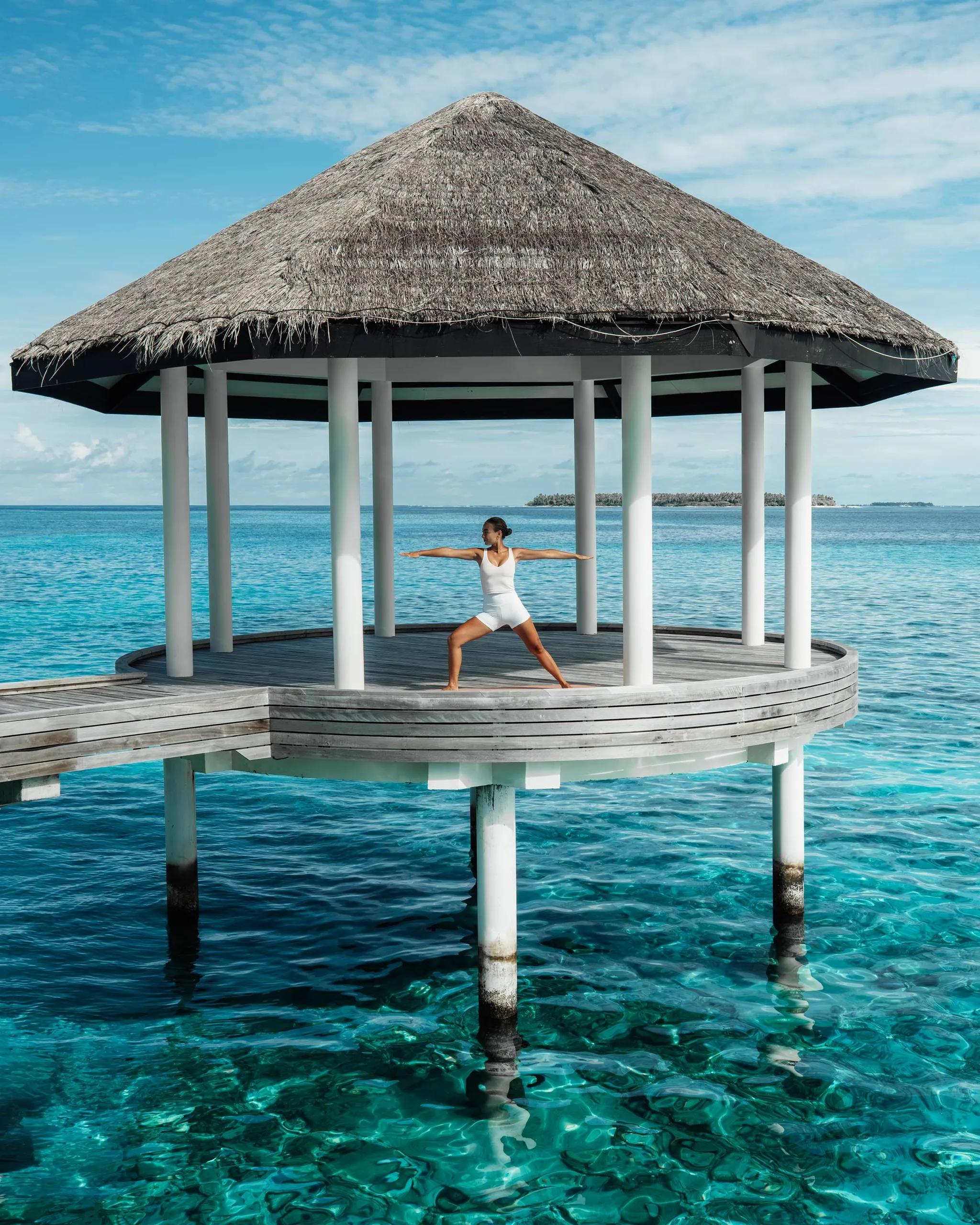 A yoga pose held on Blu Spa's overwater pavilion at Radisson Blu Resort Maldives, an outer island visible on the horizon