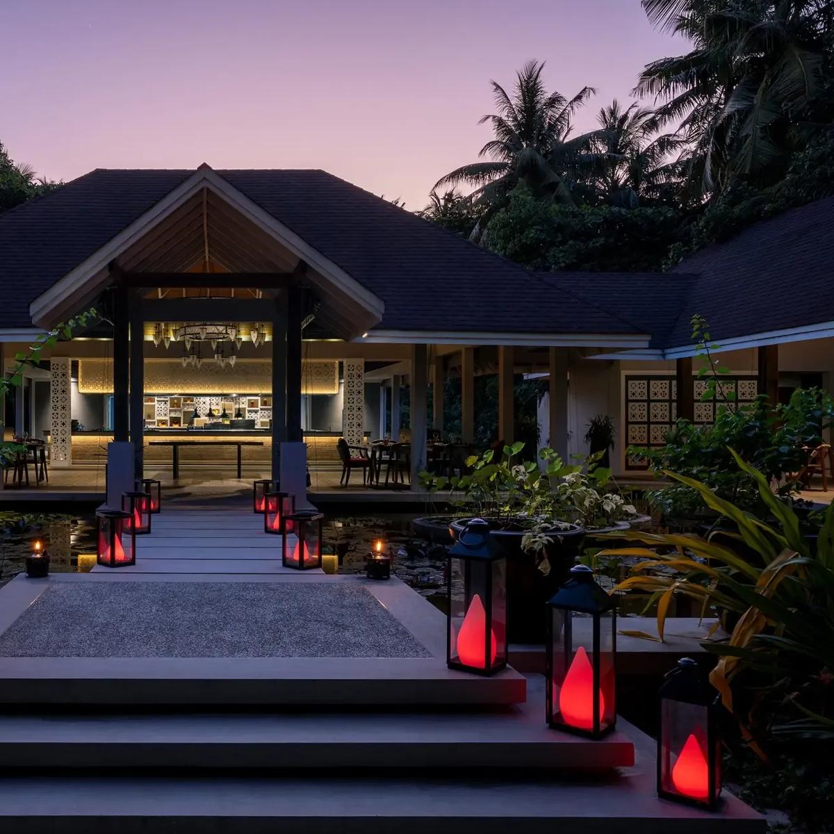 The lantern-lit walkway to Battuta at dusk, crossing a lily pond toward the open-sided dining room