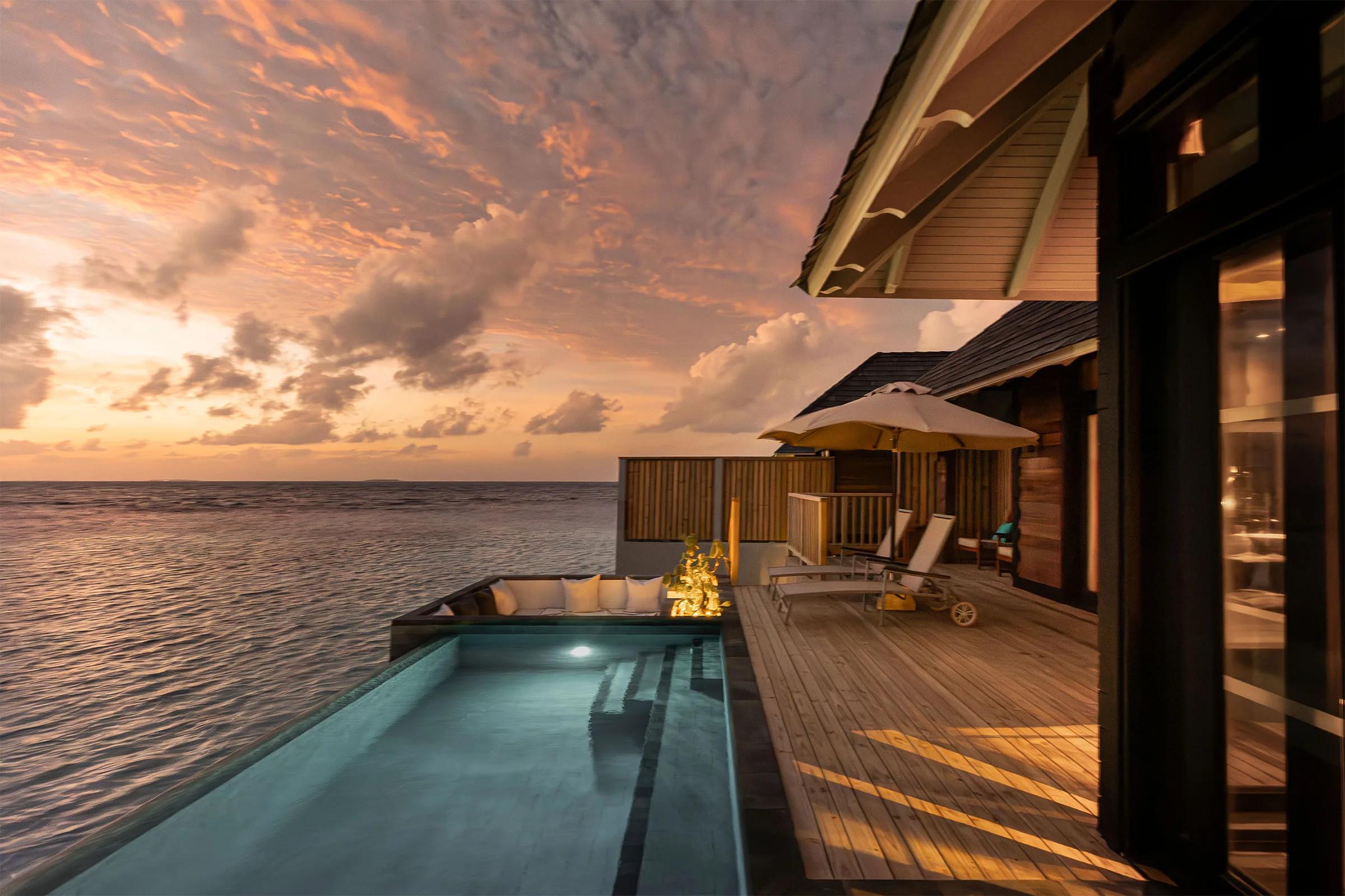 A private pool with built-in daybed seating at a Sunset Horizon Water Villa, lit by a pink and orange sunset over open water