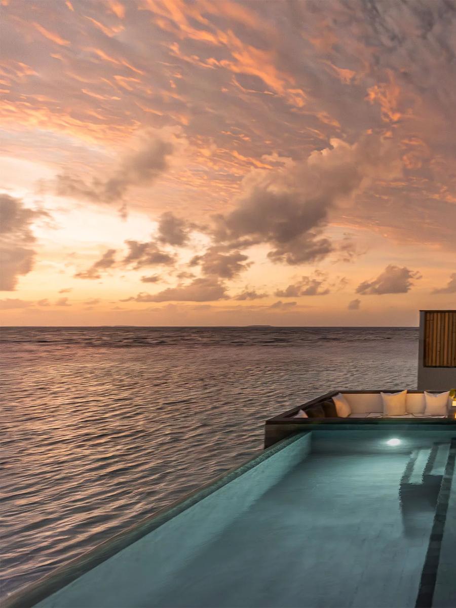 A private pool with built-in daybed seating at a Sunset Horizon Water Villa, lit by a pink and orange sunset over open water