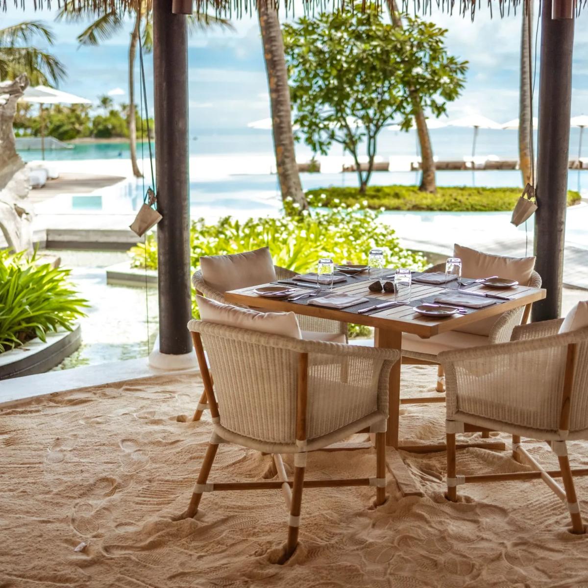 Table set for four under a thatched pergola on the sand, overlooking the pool and lagoon