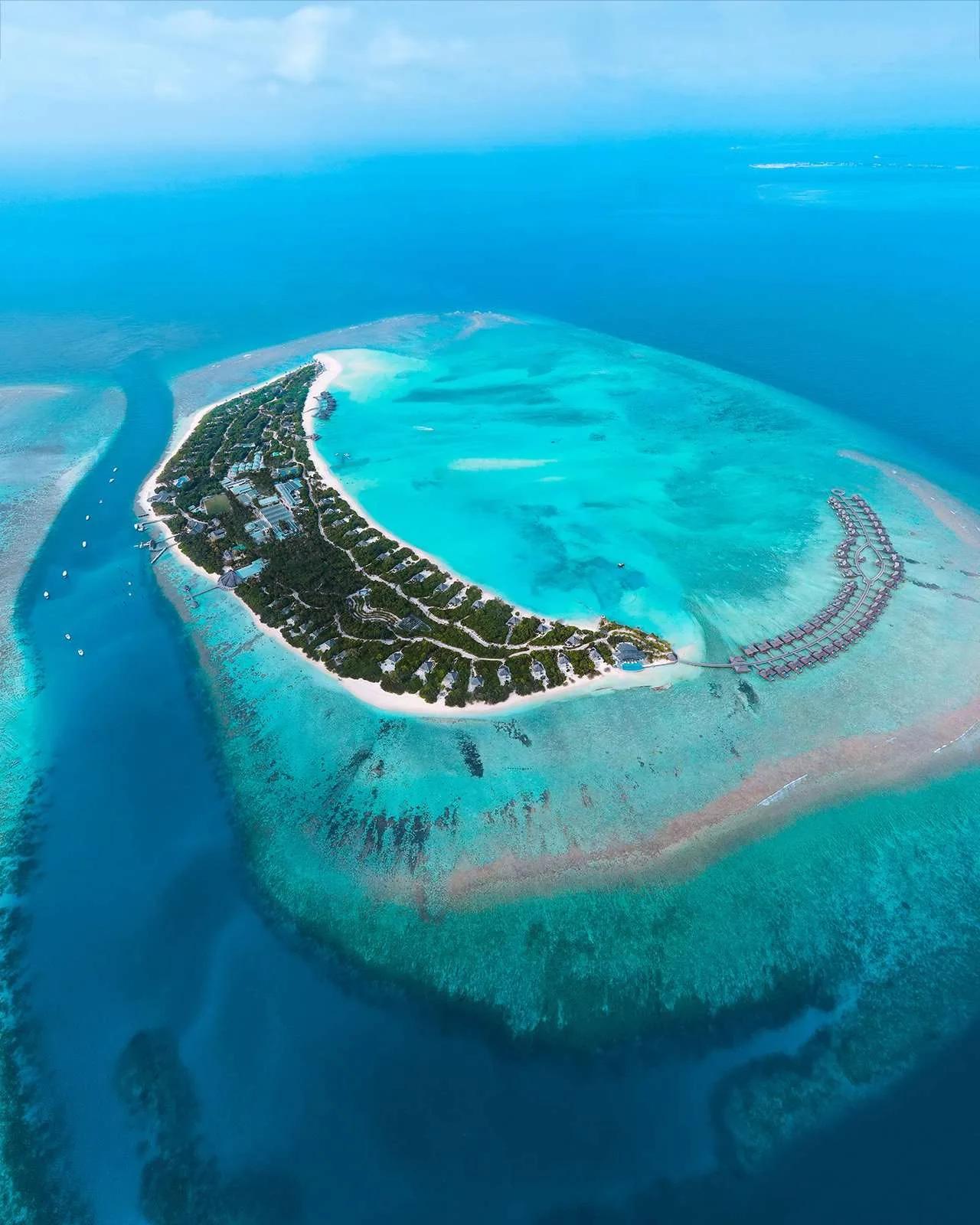 Aerial view of Dhonakulhi Island, showing Hideaway Beach Resort & Spa's full crescent shape and overwater villas at its eastern tip