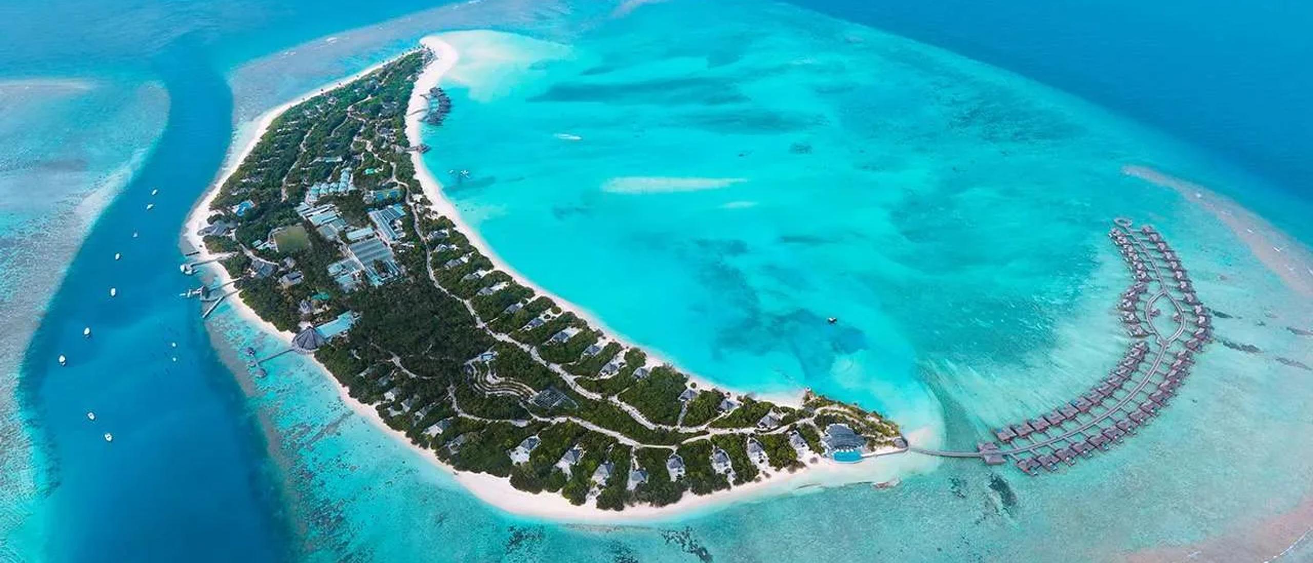 Aerial view of Dhonakulhi Island, showing Hideaway Beach Resort & Spa's full crescent shape and overwater villas at its eastern tip