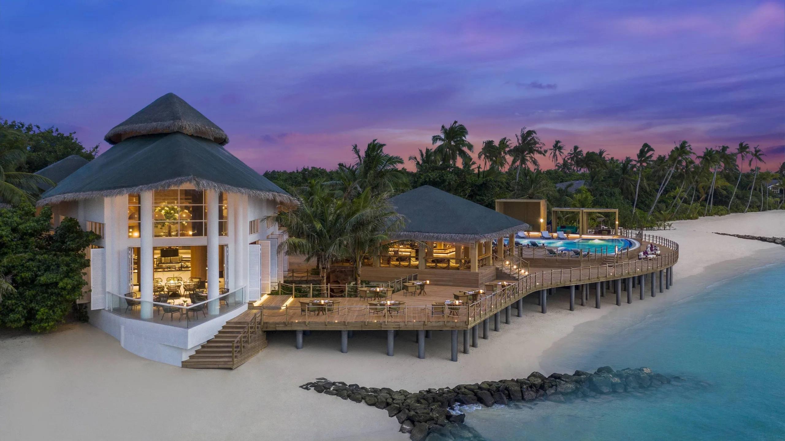 Aailaa restaurant at dusk, its thatched dining pavilion and pool deck lit up beside the beach