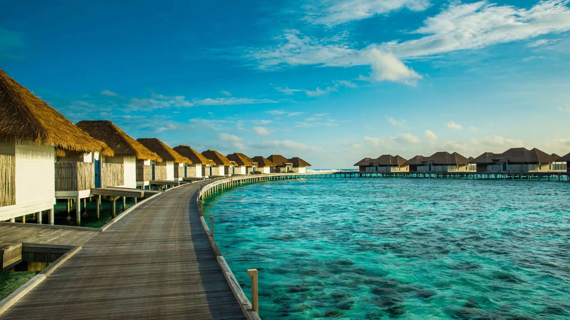 Wooden boardwalk curving between thatched-roof overwater suites toward the open lagoon under a blue sky